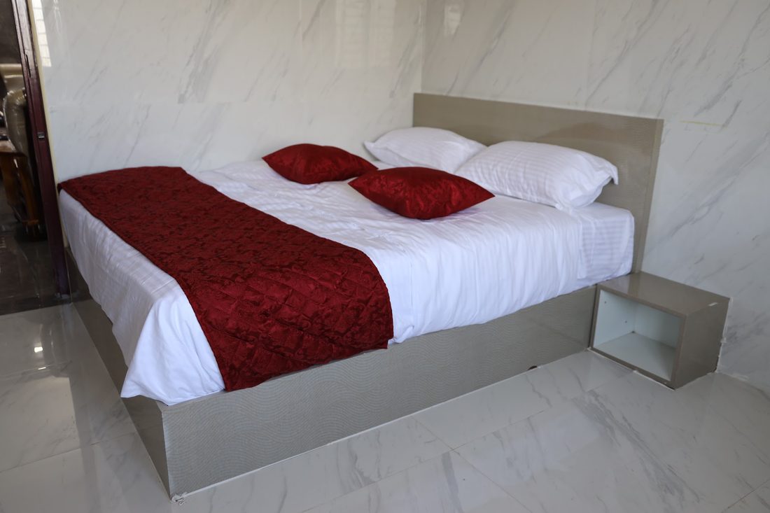 A studio unit: a double bed with a maroon runner against a marble-finish wall, with a window and a bedside table.