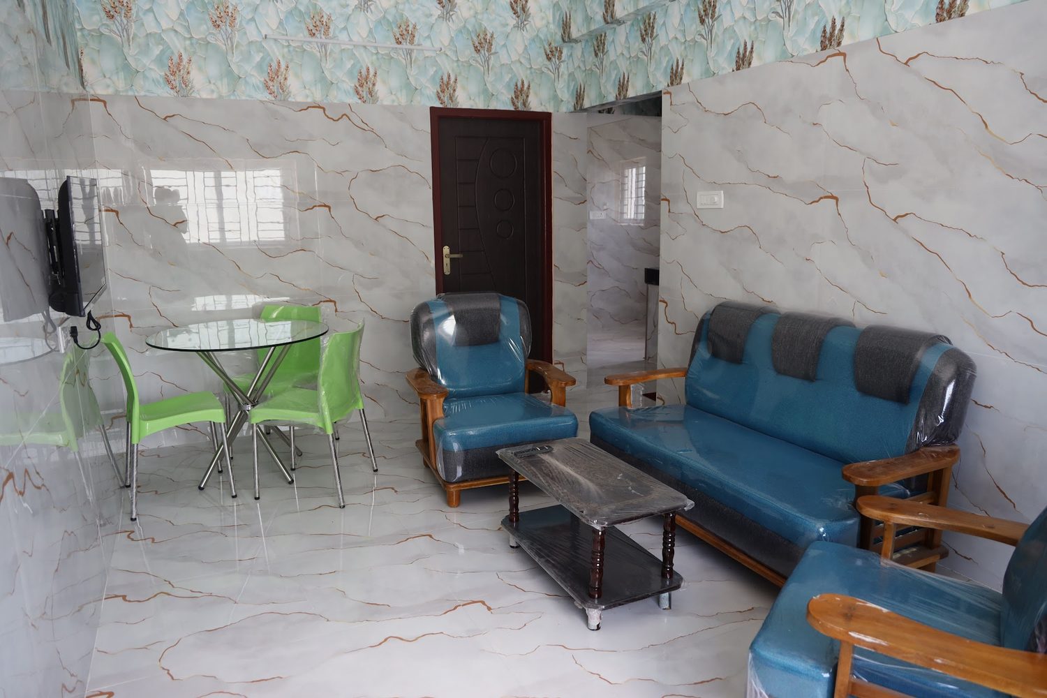 A one-bedroom apartment's living room with a teal sofa set, a low table, a round dining table with chairs and a wall-mounted television.