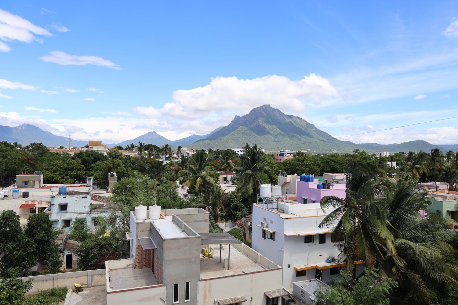 The view from an upper-floor balcony: rooftops and coconut palms in the foreground, and a broad rocky hill under a blue sky with white cloud.